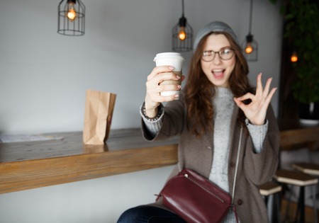 Happy young woman holding cup of coffee and showing ok sign in cafeの写真素材