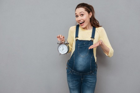 Photo of pregnant happy woman holding clock standing isolated over grey wall. Looking at camera.の写真素材
