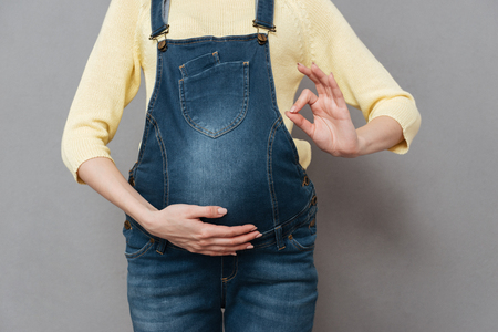 Cropped image of pregnant lady posing isolated over grey wall and make okay gesture.の写真素材