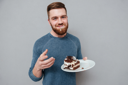 Happy smiling bearded man eating chocolate cake isolated on gray backgroundの写真素材