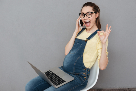 Photo of pregnant happy lady wearing glasses sitting isolated over grey wall while using laptop computer and talking by phone. Looking at camera make okay gesture.の写真素材