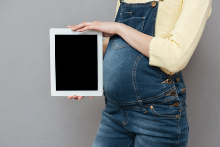 Cropped photo of pregnant woman wearing glasses standing isolated over grey wall while showing display of tablet computer.の写真素材