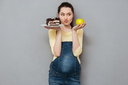 Photo of pregnant lady standing isolated over grey wall while choosing between sweet cake and apple. Looking aside.の写真素材