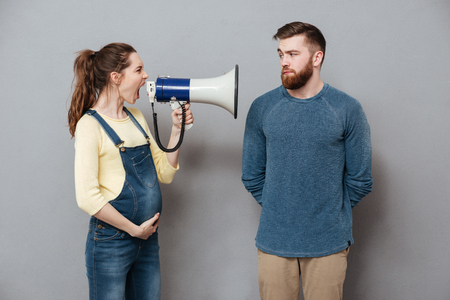 Image of pregnant screaming woman holding loudspeaker standing near serious man isolated over grey wall.の写真素材