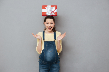 Image of pregnant happy lady standing isolated over grey wall while holding gift on head. Looking at camera.の写真素材