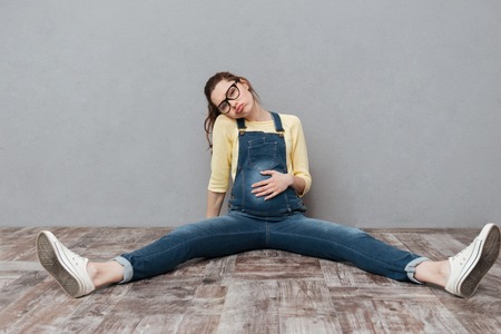 Image of pregnant tired woman wearing glasses sitting isolated over grey wall. Looking at camera.の写真素材