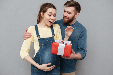 Portrait of a happy cheerful husband giving present box to his pregnant wife isolated over gray backgroundの写真素材