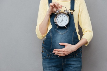Cropped photo of pregnant woman holding clock standing isolated over grey wall.の写真素材