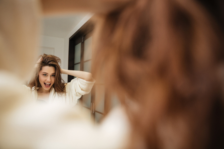Cheerful young woman in bathrobe touching her hair and having fun at bathroomの写真素材