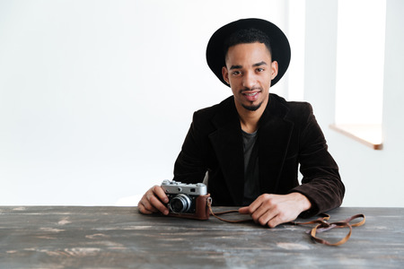 Smiling African man in suit which sitting by the table with retro camera and looking at the cameraの写真素材