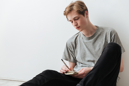 Young Man in jeans and t-shirt sitting on the floor in studio and writing something in the notebook over gray backgroundの写真素材