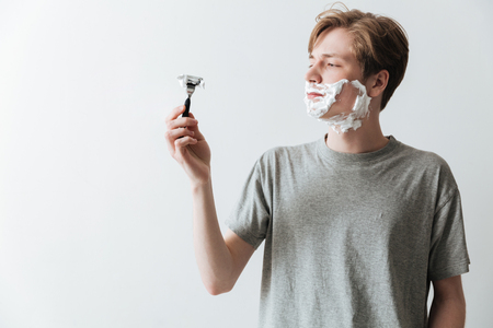 Young man in t-shirt and shaving foam looking at the razor over gray backgroundの写真素材