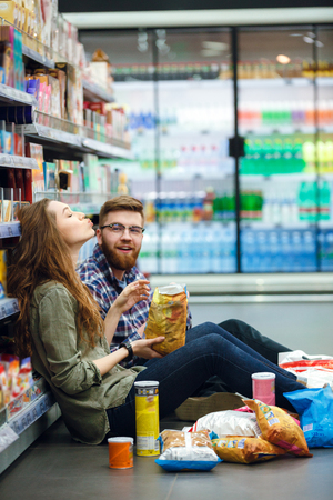 Portrait of a young funny hungry couple sitting on the supermarket floor and eating snacksの写真素材