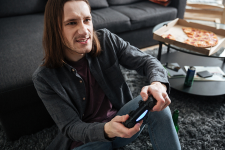 Image of young concentrated man sitting at home indoors play games with joystick. Looking aside.