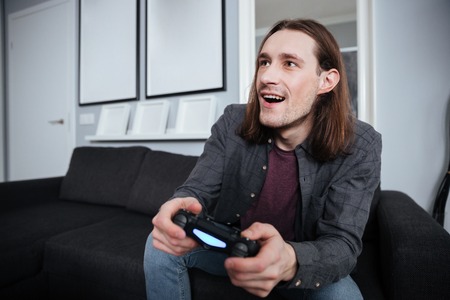 Image of young happy man gamer sitting at home indoors and play games with joystick. Looking aside.の写真素材