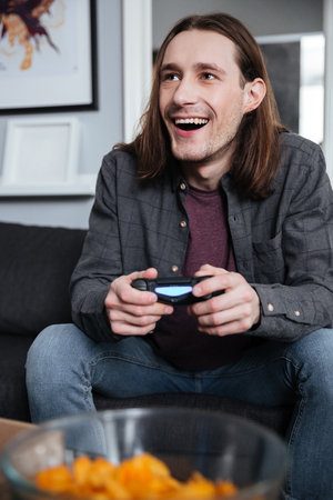 Image of young smiling man gamer sitting at home indoors and play games with joystick. Looking...
