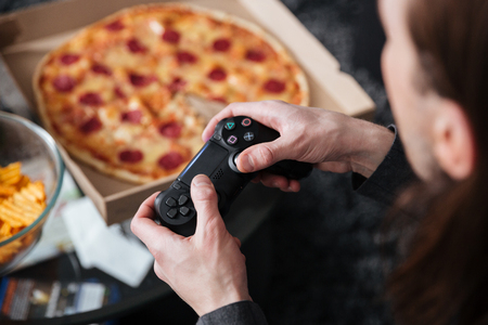 Cropped photo of young man gamer sitting at home indoors and play games with joystick near pizza and...