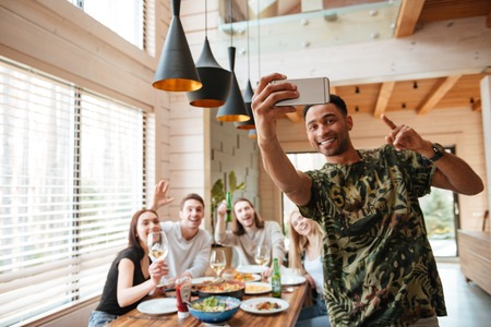 Cheerful african young man taking selfie with his friends at the table at home