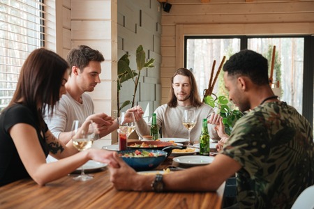 Side view picture of a group of friends praying with eyes closed over dinner table at home