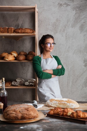 Picture of concentrated young lady baker standing with arms crossed at bakery near bread. Looking aside.の写真素材
