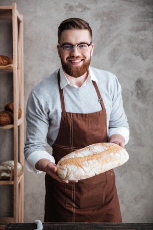 Image of cheerful young man baker standing at bakery holding bread. Looking at camera.の写真素材