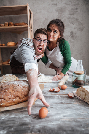 Young man and shocked woman trying to catch egg on table with flour in grey kitchenの写真素材