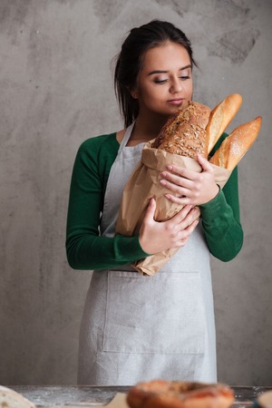 Picture of young happy lady baker standing and holding bread. Looking aside.