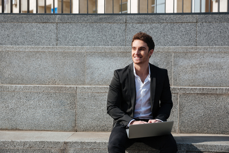 Image of happy young businessman sitting outdoors. Using laptop computer. Looking aside.の写真素材