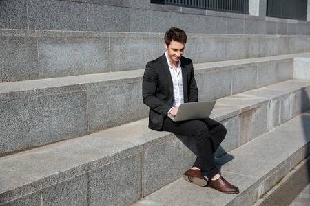 Photo of smiling young businessman sitting outdoors. Using laptop computer. Looking aside.の写真素材