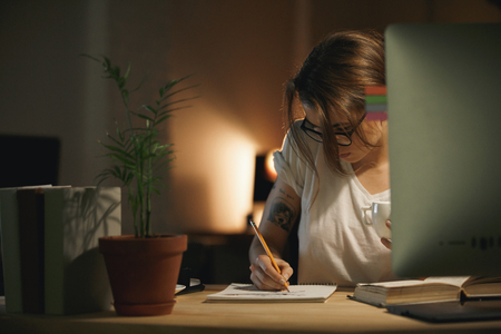 Image of concentrated young woman designer sitting indoors at night writing notes using computer. Looking aside.の写真素材