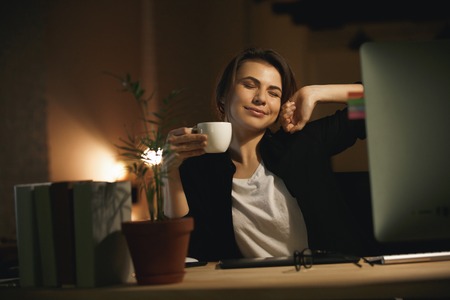 Photo of smiling young woman designer stretching indoors at night using computer and drinking coffee.の写真素材
