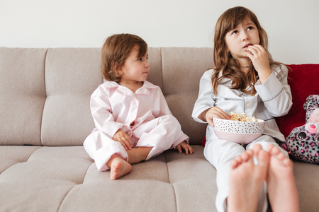 Younger sister looking at elder sister while she eating popcorn in living roomの写真素材