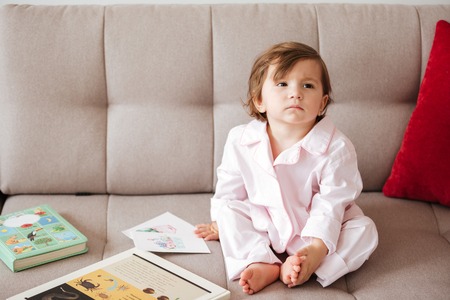 Pensive child wearing pajamas sitting on big sofa near postcards in living roomの写真素材