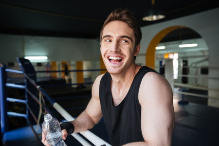 Side view of laughing boxer relaxing in boxing ring and drinking water in gymの写真素材