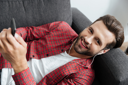Image of cheerful young man lies on sofa indoors at home while listening music with earphones. Looking at camera chatting by phone.の写真素材