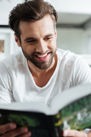 Picture of young smiling man sitting on sofa indoors at home while reading book. Looking aside.
