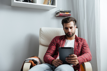 Photo of concentrated young bearded man sitting at home indoors while using tablet computer. Looking aside.の写真素材