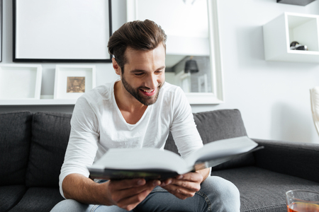 Image of young cheerful man sitting on sofa indoors at home while reading book. Looking aside.の写真素材