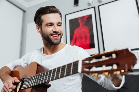 Photo of young happy man sitting on sofa indoors at home while playing on guitar. Looking aside.の写真素材