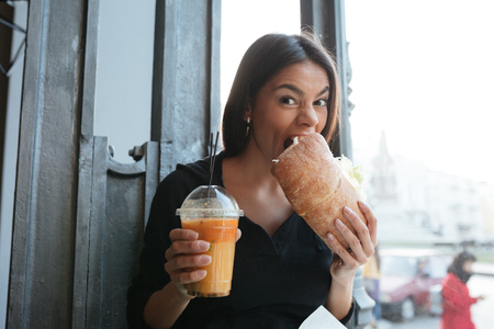 Photo of happy young woman sitting in cafe drinking juice and eating sandwich. Looking at camera.の写真素材