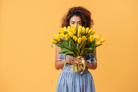Pretty African woman in blue dress hiding behind the bouquet of flowers over yellow backgroundの写真素材