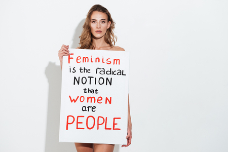 Picture of naked young caucasian lady posing over white wall holding blank with text about feminism. Looking at camera.の写真素材