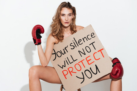 Pretty Naked woman in boxing gloves sitting on chair with broadsheet and looking at the camera over grey background.Your silence will not protect youの写真素材