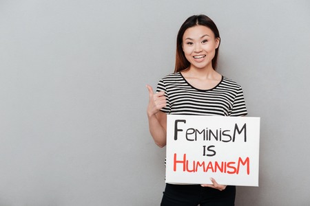 Image of young happy woman standing over grey wall showing thumbs up. Looking at camera holding blank with text about feminism.の写真素材