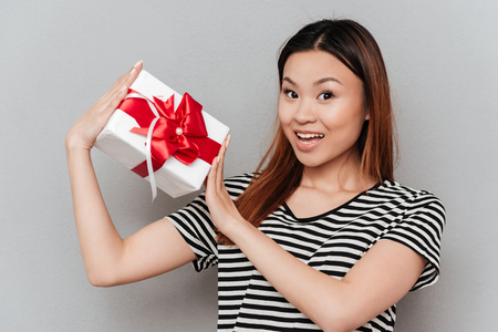 Photo of happy young woman standing over grey wall. Looking at camera holding gift.の写真素材