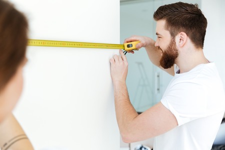 Picture of young cheerful loving couple standing indoors in new flat and make repair. Looking aside.