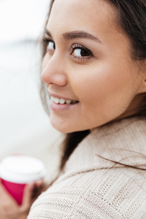 Image of cheerful young caucasian lady walking outdoors at beach wearing warm jacket drinking coffee. Looking at camera.の写真素材