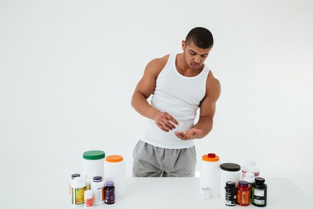 Picture of young sportsman standing over white background holding vitamins and sport pills. Looking aside.の写真素材