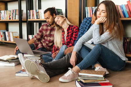 Picture of young tired students sitting in library on floor using laptop computer and reading books. Looking aside.の写真素材
