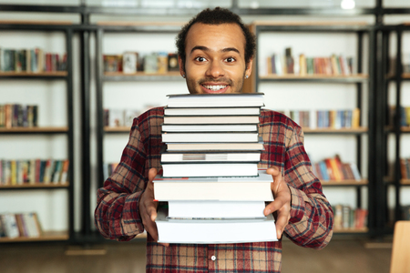 Picture of young happy african man student standing in library with a lot of books. Looking at camera.の写真素材